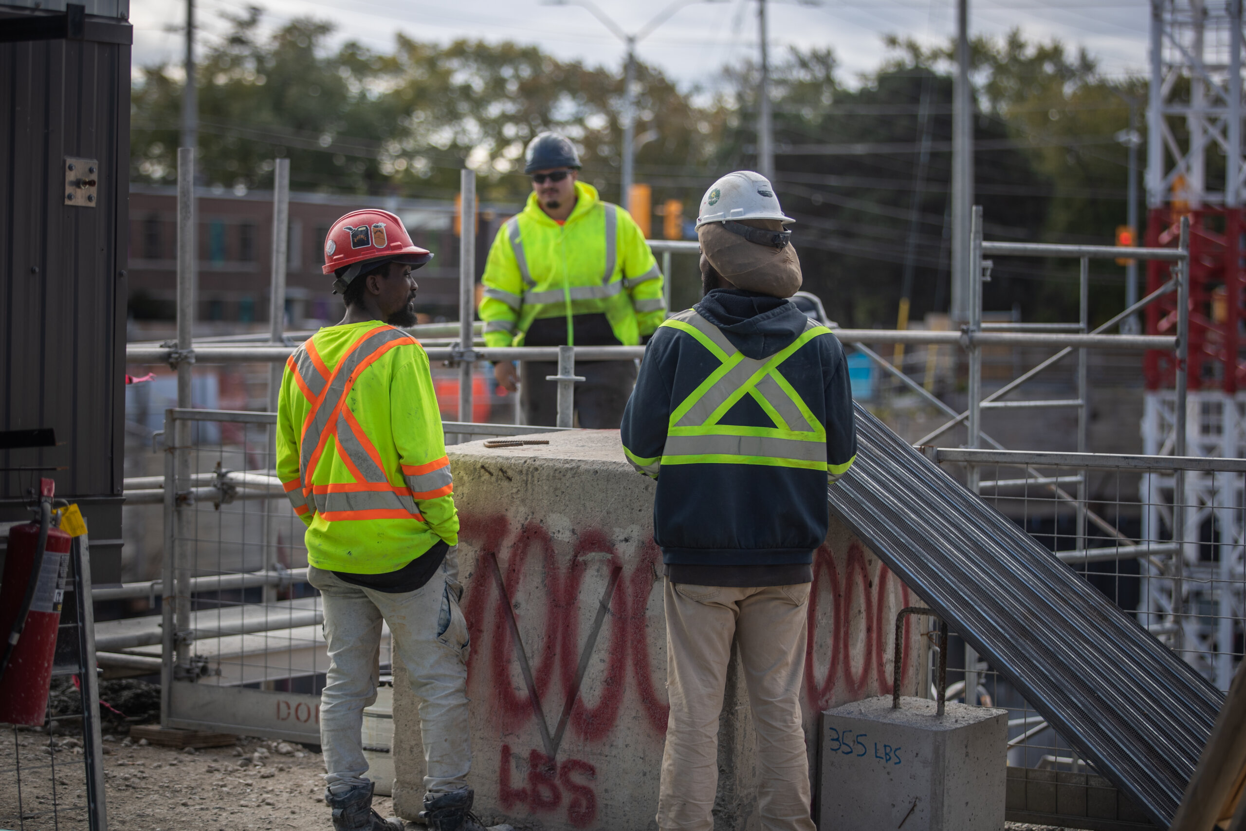 Three construction workers in high-visibility vests and hard hats standing at a jobsite near rebar cages and a concrete block marked 700 pounds.
