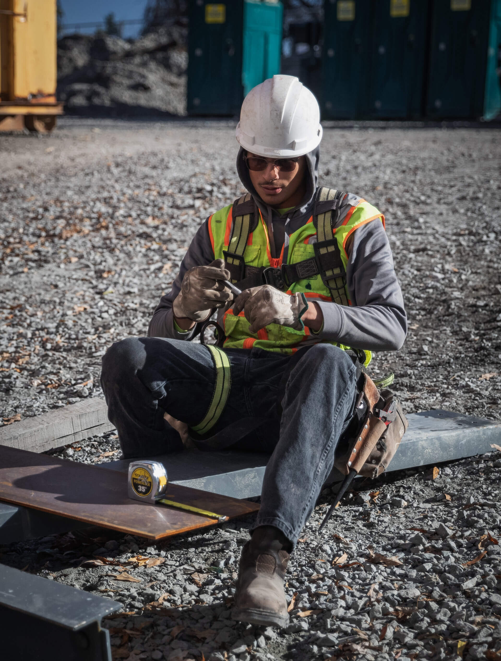 Worker in white hard hat and high-vis vest sits on gravel job site, inspecting small tool held in gloved hands