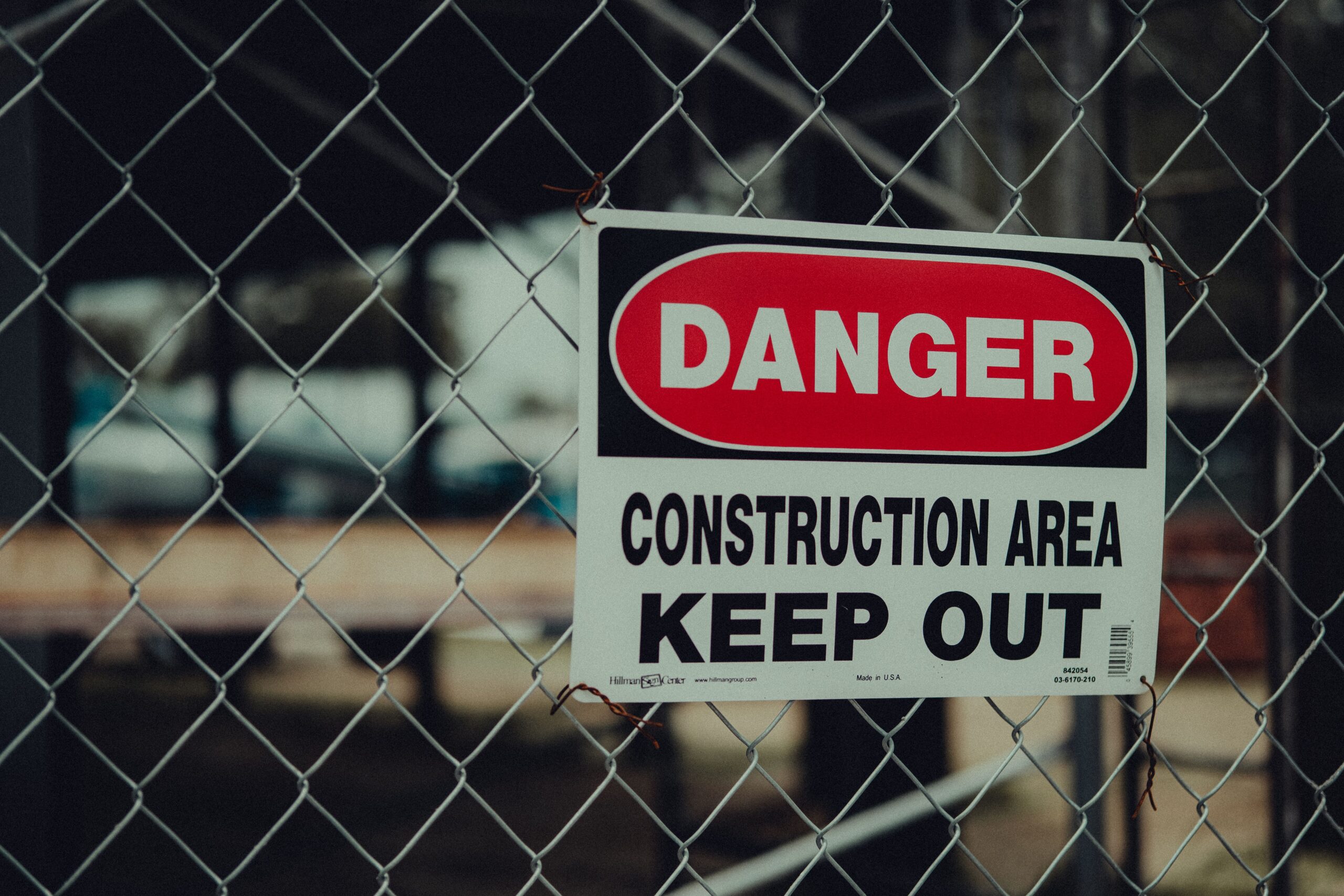 Red and white Danger sign reading Construction Area Keep Out wired to a chain-link fence, with a construction site out of focus behind it.